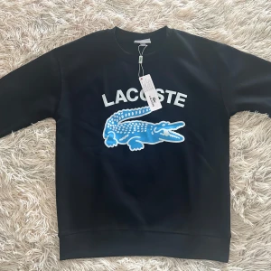 Svart sweatshirt från Lacoste - Snygg svart sweatshirt från Lacoste med det ikoniska krokodilmärket i blått och vitt på framsidan. Tröjan har en klassisk rund hals och långa ärmar. Perfekt för en stilren och avslappnad look. Tröjan är i storlek M men sitter mer som Storlek S/XS. Den säljs då den är för liten för mig. Tröjan är helt ny och aldrig använd.