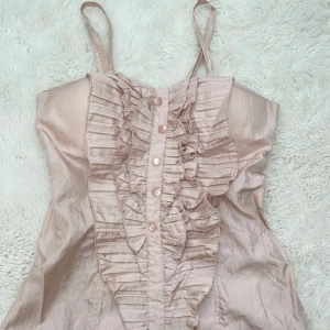 Rosa ärmlös blus med volangdetaljer - Säljer en söt rosa blus med smala axelband och volangdetaljer framtill. Blusen har knappar och ett glansigt material som ger en elegant touch. Perfekt för en somrig look!