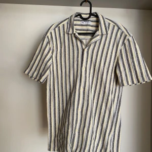 Randig kortärmad skjorta från Zara - Säljer en stilren randig skjorta från Zara i storlek L. Skjortan har korta ärmar och ett klassiskt kragdesign. Färgerna är en mix av svart, vit och gul, vilket ger en fräsch look. Perfekt för varmare dagar!