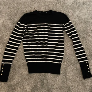 Svart och vit randig tröja med knappar - Snygg svart tröja med vita ränder och dekorativa knappar vid ärmsluten. Perfekt för en stilren look. Tröjan har långa ärmar och en klassisk passform.