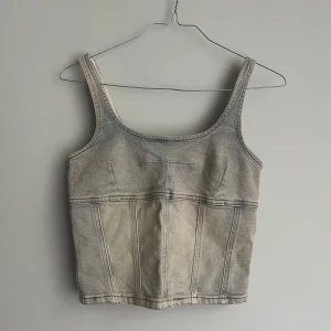 Grå jeanskorsett från Collusion - Säljer en snygg grå jeanskorsett från Collusion med dragkedja fram. Den har smala axelband och en stilren design som passar perfekt till både vardag och fest. Tillverkad i Turkiet.