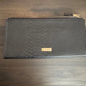 Svart plånbok från Bianca Ingrosso - Snygg svart plånbok med ormskinnsmönster från Bianca Ingrosso. Den har en praktisk dragkedja och en elegant guldlogga på framsidan. Perfekt för att hålla ordning på dina kort och kontanter.