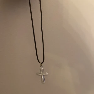 Halsband med korshänge - Snyggt halsband med ett korshänge i silverfärg. Hänget är dekorerat med små glittrande stenar och hänger på en svart rem. Perfekt för att ge en stilren touch till din outfit.