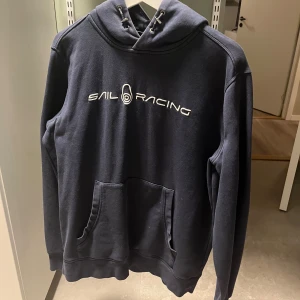Mörkblå hoodie från Sail Racing - Säljer en mörkblå hoodie från Sail Racing med vit logga på bröstet. Tröjan har en klassisk känguruficka och justerbar huva med snörning. Perfekt för en avslappnad stil.