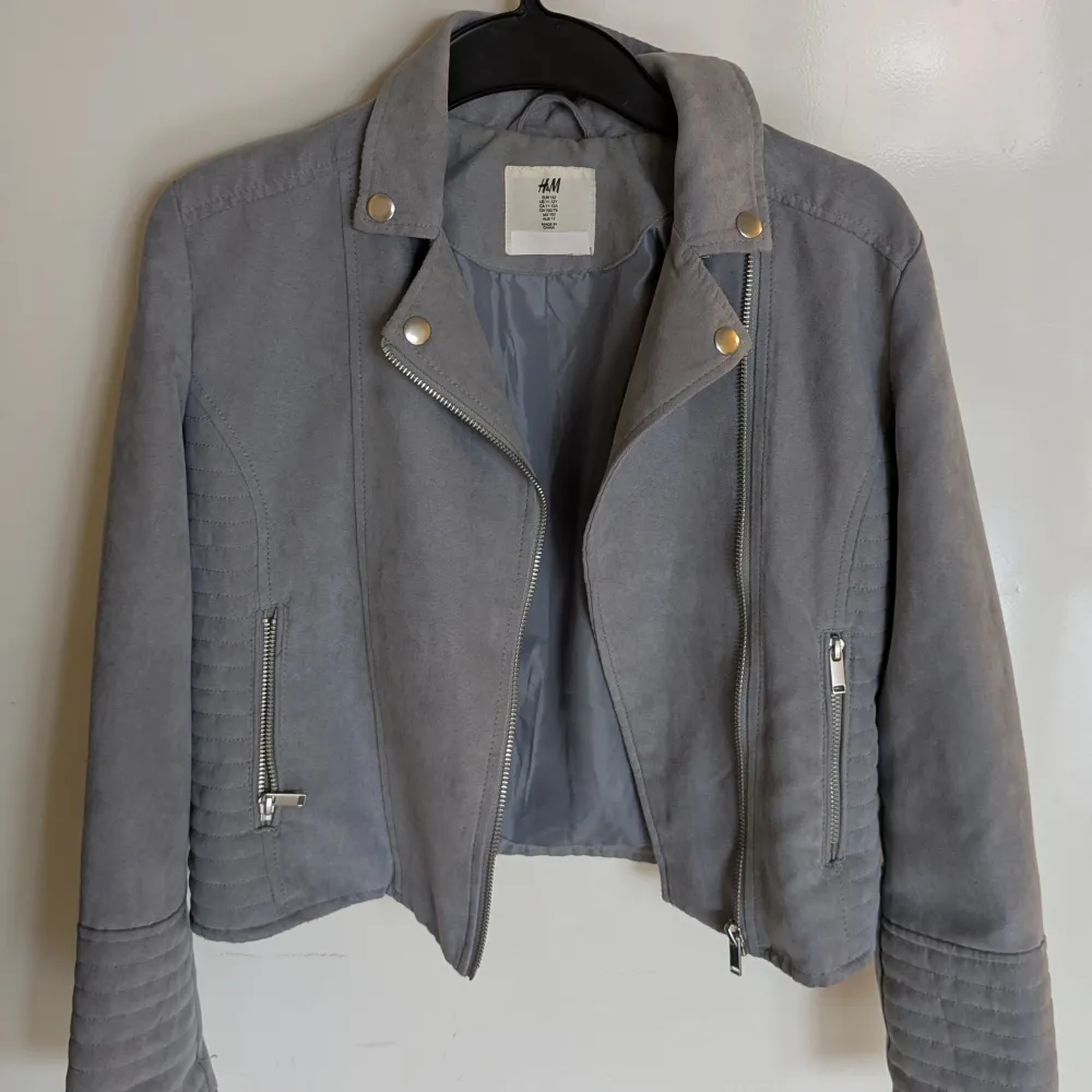 Snygg grå jacka från H&M med dragkedja och stilrena detaljer. Jackan har en klassisk bikerstil med långa ärmar och silverfärgade knappar. Perfekt för en cool och avslappnad look. 100% Polyester. Tvättas i 40 grader. Min dotters namn är tillfälligt över klistrat med ett vitt klistermärke. Jag kommer att stryka över hennes namn med svart textil penna när jackan säljs.. Takit.