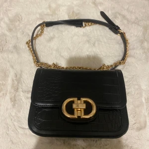 Svart axelväska med gulddetaljer - Jättesöt liten miniväska, svart axelväska med krokodilpräglat mönster och guldfärgad kedja. Väskan har en elegant guldfärgad spänne framtill och justerbar axelrem. Perfekt för att ge en lyxig touch till din outfit. 