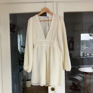 Vit kortklänning med prickigt mönster - Säljer en söt vit kortklänning med diskret prickigt mönster. Klänningen har långa ärmar och en v-ringad design som ger en elegant touch. Perfekt för sommarens festligheter!