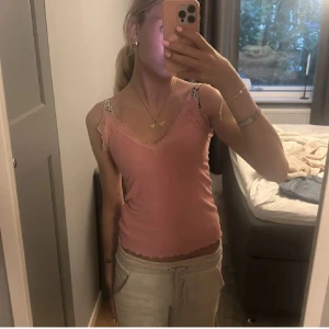 Rosa linne med spetsdetaljer - Säljer ett sött rosa linne med spetsdetaljer vid axlarna och längs kanterna. Linnet har en v-ringad design och är perfekt för varma dagar.