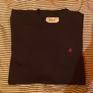 Svart t-shirt från Replay - Snygg svart t-shirt från Replay med ett litet rött broderat 'R' på bröstet. Perfekt för en stilren look. Gjord i mjukt material för extra komfort.