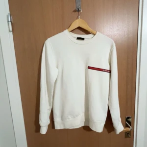 Vit sweatshirt från Prada - Säljer en stilren vit sweatshirt från Prada med rund halsringning och en diskret röd logodetalj på bröstet. Tröjan har långa ärmar och är tillverkad i mjukt material som känns skönt mot huden. Perfekt för en clean och lyxig look.