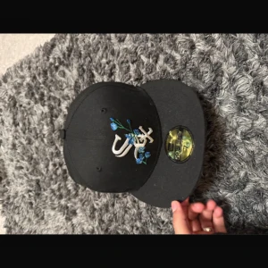 Svart keps från New Era med broderad design - Säljer en svart keps från New Era 59FIFTY med broderad vit och blå design framtill. Kepsen har en ljusblå undersida på skärmen och klassisk passform. Perfekt för dig som vill ha en stilren och unik accessoar.