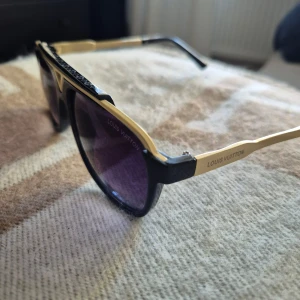 Solglasögon guld svarta - Snygga solglasögon med svarta bågar och guldfärgade detaljer. Glasen är mörklila och har en lätt toning. Klassisk design med en modern twist och tydlig logotyp på sidan.