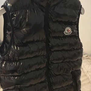 Svart dunväst från Moncler - Säljer en svart, glansig dunväst från Moncler med dragkedja framtill och klassisk Moncler-logga på bröstet. Västen har ett quiltat mönster och en unik serietecknad tvättlapp på insidan. Perfekt för lager-på-lager-stil.