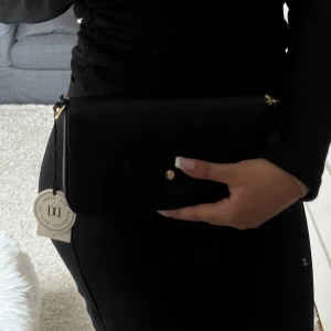 Svart axelväska med guldkedja från Don Donna ny pris 350 - Snygg svart axelväska från Don Donna med guldfärgad kedja och enkel knappstängning. Väskan har stilren design och passar perfekt till att bära det viktigaste. Märkets logga syns diskret baktill.