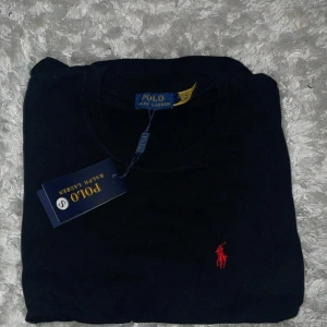 Svart t-shirt från Polo Ralph Lauren - Svart t-shirt från Polo Ralph Lauren med klassisk rund hals och röd broderad logga på bröstet. Enkel och stilren design som passar till det mesta. Perfekt basplagg för garderoben.