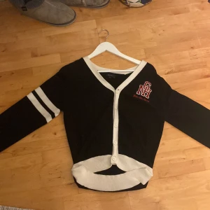 Svart kofta med vita detaljer från Gant - Svart kofta från Gant med vita ribbade kanter och knappar framtill. V-ringad modell med broderad röd och vit logga på bröstet samt två vita ränder på ena ärmen. Perfekt för en sportig och stilren look.