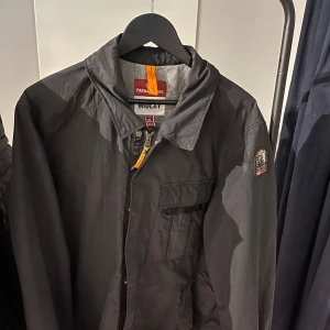 Svart jacka från Parajumpers Midlay - Säljer en svart Parajumpers Midlay jacka i storlek XL. Sitter som L Jackan dragkedja framtill och en bröstficka med knapp. På vänster ärm sitter det ikoniska PJS-märket. Perfekt för dig som gillar stilren design.