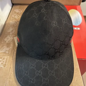 Svart keps med monogram - Säljer en svart keps med diskret GG-monogram över hela kepsen och klassiska gröna och röda ränder på sidan. Justerbar rem baktill för perfekt passform. Snygg och stilren accessoar för dig som gillar exklusiva märken.