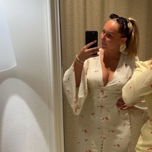 Vit blommig klänning från Adoore - Superfin vit kortklänning från Adoore med små röda blommor, v-ringning och knappar framtill. Klänningen har långa, vida ärmar och markerad midja. Perfekt för sommarens alla tillfällen! (Armarna är uppvika på mina egna bilder men ser ut som på hemsidan) Den är endast använd ett fåtal gånger och är i storlek 42 (ganska liten i storleken) 🌺🍓☀️