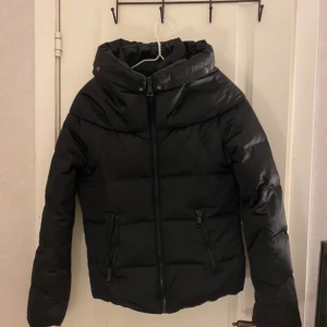 Svart pufferjacka  - Säljer en svart pufferjacka utan  huva och med dragkedja framtill. Jackan har två fickor med dragkedja och hög krage. Perfekt för kyligare dagar och har en klassisk, enkel design.