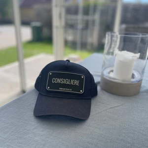 Svart keps från John Hatter & Co med Consigliere-tryck - Svart keps med nät baktill och justerbar passform. Framtill finns en stor skylt med texten 'CONSIGLIERE'. Märket John Hatter & Co syns tydligt. Perfekt accessoar för en cool och avslappnad stil.