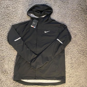 Nike vindjacka - Nike Shieldrunner vindjacka i svart med reflekterande detaljer och huva. Jackan har dragkedja framtill, två sidofickor och är tillverkad i ett vindtätt material. Perfekt för löpning eller utomhusaktiviteter när det blåser.