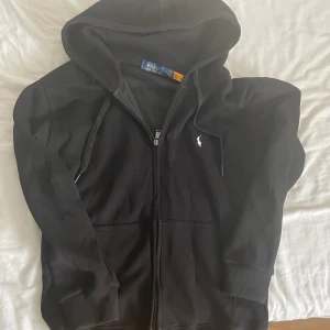 Ralph lauren kofta - Säljer en svart hoodie från Polo Ralph Lauren med dragkedja och huva. Klassisk design med vit broderad logga på bröstet och ribbade muddar. Perfekt för en avslappnad stil.