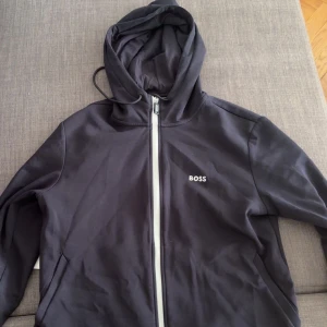 Svart hoodie med dragkedja från BOSS - Säljer en svart hoodie från BOSS med vit dragkedja framtill och huva. Tröjan har fickor på sidorna och BOSS-logga broderad på bröstet. Perfekt för en avslappnad stil. Original pris: 999kr