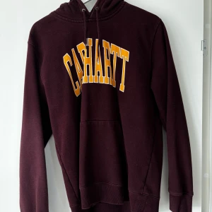 Vinröd hoodie från Carhartt - Säljer en vinröd hoodie från Carhartt med stor gul och vit logga på bröstet. Tröjan har huva med dragsko och en stor magficka. Perfekt för dig som gillar streetwear och vill ha något bekvämt och snyggt.