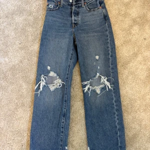 Levi's Ribcage Straight Ankle jeans med slitningar - Säljer ett par blå Levi's Ribcage Straight Ankle jeans med medelhög midja och raka ben. Jeansen har slitna detaljer och hål vid knäna för en trendig look. Klassisk femficksmodell med knappgylf och Levi's-lapp bak. PRIS GÅR ATT DISKUTERA!!