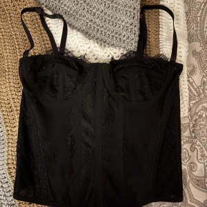 Svart spetskorsett med axelband - Snygg svart korsett med spetsdetaljer och justerbara axelband. Modellen har markerade kupor och dekorativa sömmar framtill. Perfekt för att lyfta en outfit med en trendig touch. En topp som passar perfekt för utgång med som utan kavaj! OBS. Sista bilden lånad, men så sitter den på :)