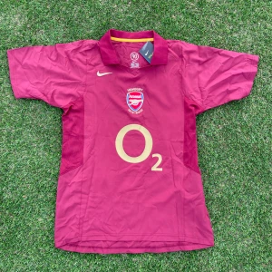 Arsenal Hemma Kit 2005/06  - Säljer en klassisk Arsenal fotbollströja i vinrött med guldiga detaljer och tryck. Tröjan har korta ärmar, krage och klubbmärke på bröstet samt Henry 14 på ryggen. Perfekt för fans av Arsenal och Thierry Henry.