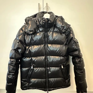 Svart pufferjacka från Moncler - Säljer en svart pufferjacka från Moncler med huva och dragkedja. Jackan har två framfickor med dragkedja och en ärmficka med Moncler-logga. Klassisk quiltad design och glansig finish.