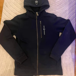 Mörkblå hoodiejacka från Sail Racing - Mörkblå hoodiejacka med dragkedja från Sail Racing. Jackan har huva med broderad logga och en bröstficka med dragkedja. Perfekt för svala dagar och enkel att matcha med det mesta.