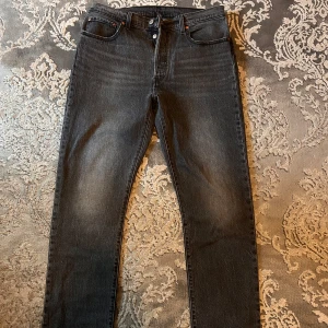 Levi's 501 svarta jeans - Klassiska svarta Levi's 501 jeans med raka ben och femficksdesign. Jeansen har knäppning framtill och subtila slitningar på låren för en snygg vintage-look. Perfekta för dig som gillar tidlös stil. Inte använda alls. Pris kan diskuteras. Storlek W 32 L 32 