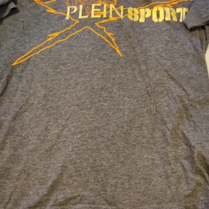 Grå t-shirt från Plein Sport med gult tryck även mera från Plein  - Säljer en grå t-shirt från Plein Sport med stort gult tryck och grafisk design framtill. T-shirten har klassisk passform och är perfekt för en sportig look. Röd tshirt och vit hoodie. 95kr/st för tshirts och 285kr för hoodie 