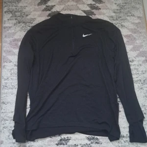 Svart långärmad träningströja från Nike - Säljer en svart långärmad träningströja från Nike Running med Dri-Fit-material. Tröjan har en liten dragkedja framtill och reflekterande detaljer på ryggen. Perfekt för löpning eller annan träning.