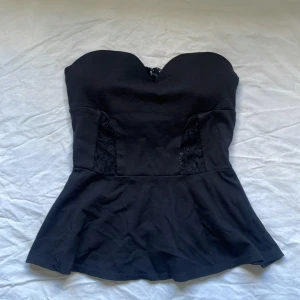 Svart axelbandslös topp från Tally Weijl - Säljer en svart axelbandslös topp från Tally Weijl med spetsdetaljer på sidorna och en dragkedja i ryggen. Toppen har en figurnära passform upptill och utsvängd nertill, perfekt för en trendig look. Inbyggd BH