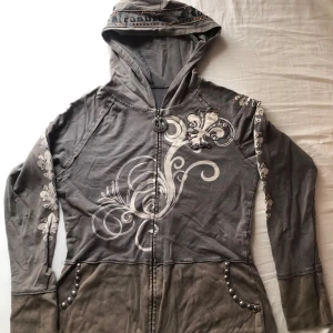 Hoodie från Treasure Rock - Säljer en grå/beige hoodie med tryck och nitar längs fickorna. Huvan har text och dekorerande stenar. (Saknar några stenar, men snygg ändå)