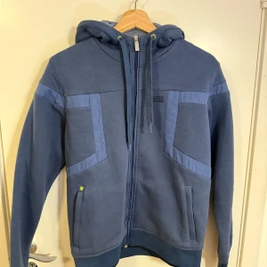 Blå Hugo Boss hoodie – helt ny! - Säljer en riktigt snygg och stilren hoodie från Hugo Boss i färgen blå. Den är aldrig använd och i helt nytt skick!  Modellen är tjock och varm, perfekt för kyligare dagar. Passar storlek S/M, så både slim och lite mer relaxed fit funkar beroende på kroppstyp.  Nypris ca 2000 kr, nu säljer jag den för 799 kr. Pris kan diskuteras vid snabb affär!  Skicka ett meddelande om du är intresserad eller vill ha fler bilder! 