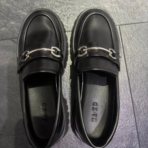 Svarta loafers med metallspänne från Na-Kd - Stilrena svarta loafers i skinn med chunky sula och silverfärgat metallspänne framtill. Klassisk design med rund tå och platt sula, perfekt för en trendig och stilren look.