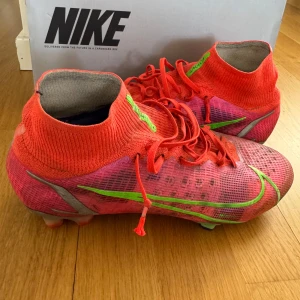 Nike Mercurial fotbollsskor i rött och neon - Säljer ett par Nike Mercurial fotbollsskor i rött med neongröna detaljer och snörning. Skorna har en stickad, strumpliknande krage för extra stöd och en platt sula med dobbar för bra grepp på planen. Perfekta för dig som vill sticka ut på fotbollsplanen! Skorna har ACC+ vilket ger en bra kontroll på bollen i alla väder!