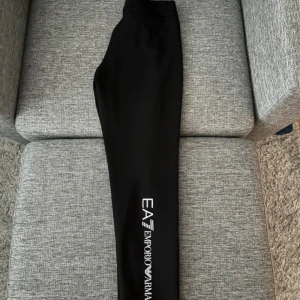 Svarta EA7 Emporio Armani mjukisbyxor - Svarta mjukisbyxor från EA7 Emporio Armani med logga tryckt längs ena benet. Byxorna har elastisk midja och smal passform, perfekta för en avslappnad stil. Materialet känns mjukt och stretchigt.