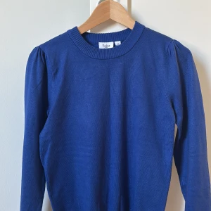 Blå stickad tröja från Saint - Snygg blå stickad tröja från Saint med rund halsringning och långa ärmar. Tröjan har diskreta puffdetaljer vid axlarna och ribbade muddar vid ärmslut och nederkant. Perfekt för lager-på-lager under kyligare dagar.