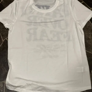 Vit t-shirt med svart text 'Faith Over Fear' - Vit t-shirt med stor svart text på ryggen: 'FAITH OVER FEAR' och bibelcitat från Psalms 118:6. Klassisk rund hals och korta ärmar. Perfekt för dig som vill bära ett tydligt statement.