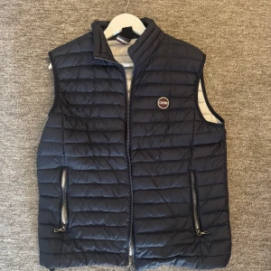 Mörkblå quiltad dunväst från Colmar - Säljer en mörkblå quiltad dunväst från Colmar med dragkedja framtill och två fickor med dragkedja. Västen har en hög krage och är lätt vadderad, perfekt för lager-på-lager under kyliga dagar.