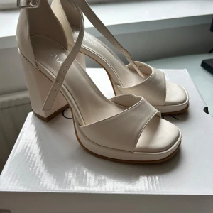 Beige sandaler med klack och ankelrem /Helt nya  - Säljer ett par helt nya  och oanvända stilrena beige sandaler med hög blockklack och justerbar ankelrem med spänne. Skorna har öppen tå och bred rem framtill, perfekt för att lyfta din outfit. Kvitto finns också.