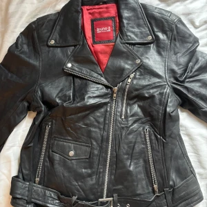Svart skinnjacka från Bruno Z - Säljer en klassisk svart skinnjacka från Bruno Z med röd insida. Jackan har sned dragkedja, flera fickor med dragkedjor och bälte i midjan. Perfekt för dig som gillar en tidlös bikerstil.