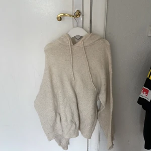 Beige stickad hoodie - Mysig beige stickad hoodie med huva och dragsko. Tröjan har långa ärmar och en avslappnad passform, perfekt för kyliga dagar. Enkel och stilren design som passar till det mesta.