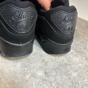 Svarta Nike Air Max sneakers - Säljer ett par helsvarta Nike Air Max sneakers med klassisk design. Skorna har mesh och syntetpaneler, snörning och en platt sula med synlig Air-dämpning. Perfekta för dig som gillar stilrena och bekväma sneakers.